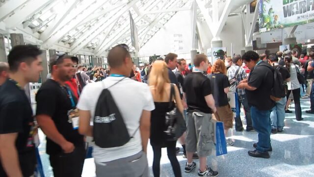 A large group of people standing in line inside a bright, spacious convention center. The attendees, wearing lanyards and carrying bags, appear to be at an event or expo. The setting suggests a bustling, lively atmosphere typical of large conventions.