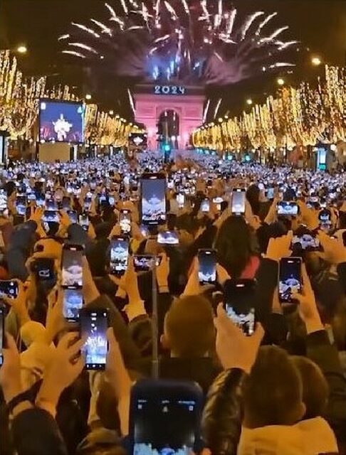 A large crowd gathered in front of the Arc de Triomphe, illuminated by decorative lights and, fireworks and lights displaying "2024." The audience is recording the event with their smartphones, capturing the spectacle.