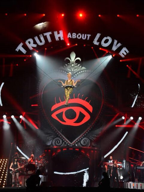 A stage performance from P!nk's "The Truth About Love" tour, featuring the singer suspended in the air with ropes, surrounded by a large illuminated heart symbol with an eye design in the center. The stage is set with vibrant red and white lights, and the words "TRUTH ABOUT LOVE" are prominently displayed above the stage.