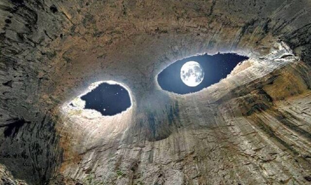 A mesmerizing view of Prohodna Cave in Bulgaria, where two large, eye-shaped openings in the cave ceiling resemble a pair of watching eyes. Through one of the openings, a bright full moon is visible against a starry night sky.