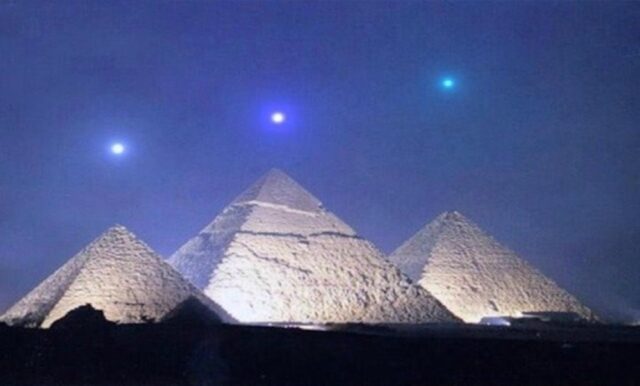 The Pyramids of Giza at night, illuminated against the night sky. Above the pyramids, three bright lights are visible, aligned in a pattern resembling Orion's Belt. The lights are blue and white, creating a striking visual alignment with the pyramids below.