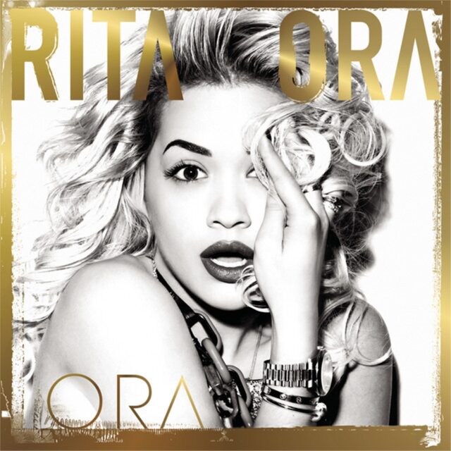 The music album cover of "Ora" (2012) by Rita Ora. In the black-and-white photo, Rita Ora is covering her left eye with her hand and hair while wearing large chains, bracelets, and rings. Her name, "RITA ORA," is displayed in bold gold letters at the top, and the album title, "ORA," is also written in gold at the bottom.