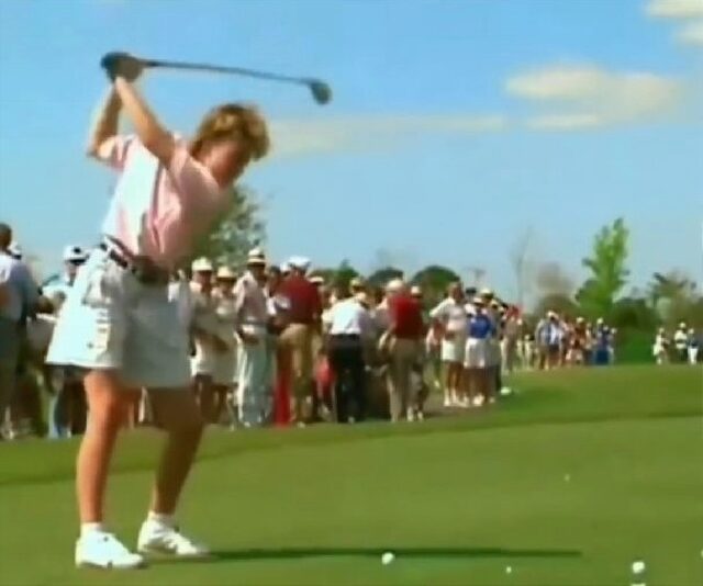 A golfer is in mid-swing on a golf course, with a crowd of spectators standing closely in front of her, observing the action. The spectators are positioned in a way that puts them in potential danger if a shot goes astray. The scene is set on a sunny day with a clear blue sky.