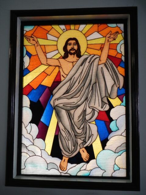 A stained glass window depicting Jesus with his arms raised, forming the shape of the letter "Y", rising amidst clouds with a radiant halo resembling the sun behind his head. His eyes are portrayed differently, adding a unique detail to the artwork. This window is located in the parish church Santuario de San Jose in East Greenhills, Mandaluyong, Philippines.