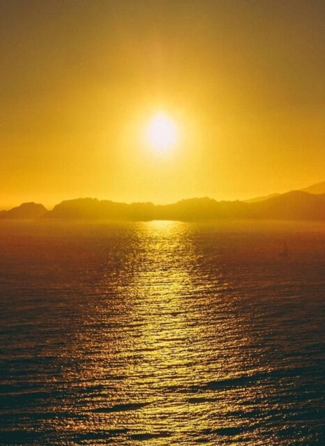 A golden sunset over a calm body of water, with the sun reflecting on the water's surface and distant hills silhouetted against the sky.