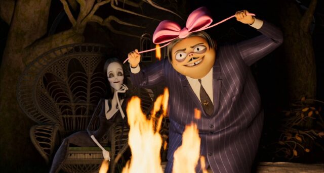 Gomez Addams, wearing a pinstripe suit and a large pink bow on his head, stands energetically by a campfire. Behind him, Morticia Addams, with her signature pale skin and dark hair, sits calmly in a wicker chair, observing the scene. The background features dark, twisted tree branches.