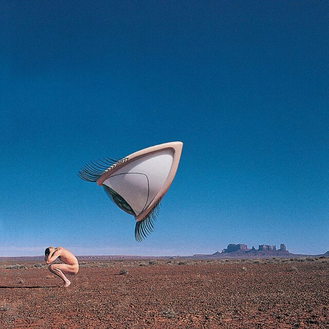 Album cover for "Bury the Hatchet by The Cranberries" (1999), featuring a surreal image of a giant, floating eye with long lashes, staring at the crouching human figure below in a stalker-like manner. The scene takes place in a barren desert landscape with distant rock formations under a clear blue sky.