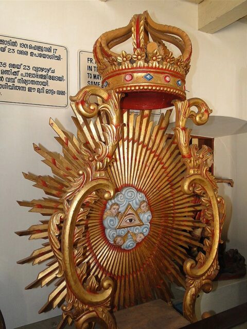 A golden altar piece from the old Catholic cathedral in Pala, Kerala, India, featuring the Eye of Providence within a triangle surrounded by clouds and cherubic faces. Rays of light extend outward from the central emblem, with a decorative crown perched on top, symbolizing divine authority.