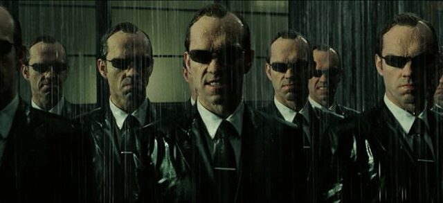 A still from "The Matrix" depicting multiple Agent Smiths standing in the rain, showcasing the concept of herd mentality as all figures appear identical, symbolizing the loss of individuality.