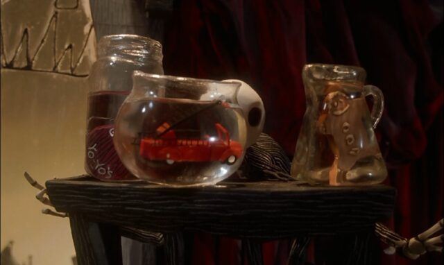 Jack Skellington peers into a bowl containing a toy fire truck floating in water. The bowl is positioned on a small table next to two jars with water, one containing a yo-yo labeled "yo-yos" and the other holding a gingerbread man.