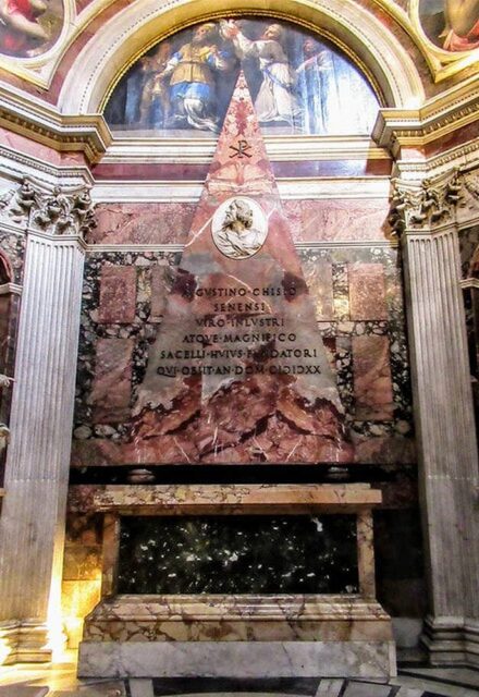 The pyramid-shaped tomb of Agostino Chigi inside the Chigi Chapel in Santa Maria del Popolo, Rome. The tomb is made of red marble and features a prominent Christian symbol, "Xpistos," at the top, which is a symbol of Jesus as the letter "P" on the cross represented by the letter "X." Below the symbol is a medallion with a portrait of Agostino Chigi, and an inscription in Latin is engraved on the front of the pyramid. The tomb is flanked by two marble columns and is set against an ornate backdrop of classical architecture and religious artwork.