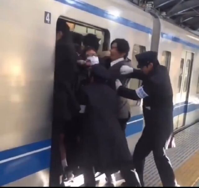 Train conductors pushing passengers into an overcrowded train to ensure the doors can close. The scene depicts a common rush hour situation in some cities, highlighting the extreme measures taken to accommodate the high number of commuters.