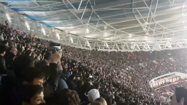 A crowded stadium filled with spectators performing the Mexican wave, which is moving towards the camera. Many people in the audience are holding up their phones to record the event. The stadium's roof structure is visible with bright lighting illuminating the scene.