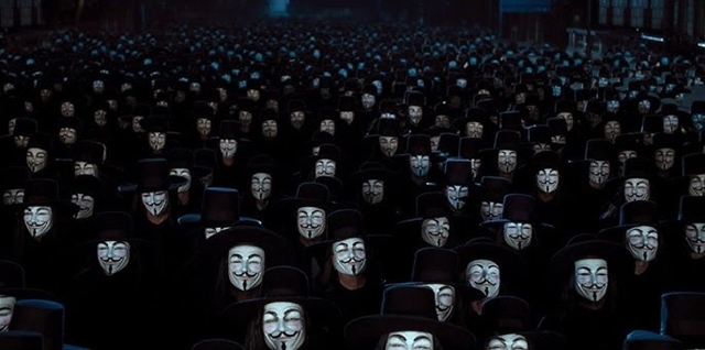 A large crowd of people wearing Guy Fawkes masks and black hats, inspired by the film 'V for Vendetta,' standing closely together in a nighttime setting. The masks, with their distinctive mustache and goatee, create a sea of identical faces.